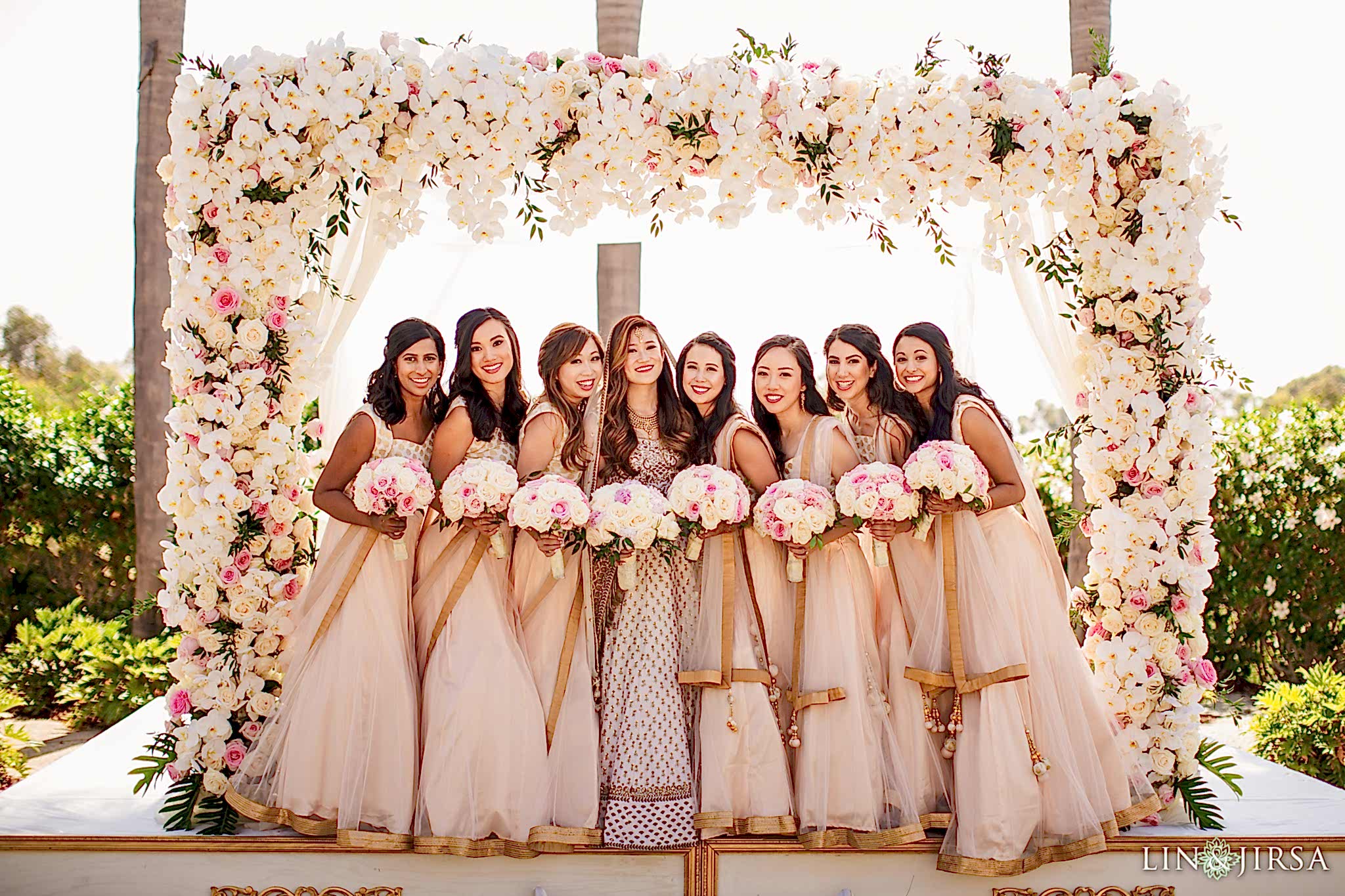 Bridesmaids posing with white and pink rose flower bouquets with the bride, under a white rose archway and palm trees, near the beach for Lin and Jirsa Los Angeles at the Park Hyatt Aviara Resort in Carlsbad, California Seven bridesmaids posing with white and pink rose flower bouquets in their hands, with the bride, under a white rose archway and palm trees, near the beach for Lin and Jirsa Los Angeles at the Park Hyatt Aviara Resort in Carlsbad, California