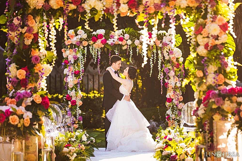 A wedding photograph shot by Chris Lin and Pye Jirsa of Lin and Jirsa Los Angeles with the bride and groom holding each other under ornate wedding arches that are draped with orange, pink, yellow and red roses and wisteria flowers, planned by Christina of Lavish Weddings at the San Diego Museum of Art , in San Diego, California A wedding ceremony photograph shot by Lin and Jirsa Los Angeles with the bride and groom holding each other under ornate wedding arches that are draped with orange, pink, yellow and red roses and wisteria flowers, planned by Christina of Lavish Weddings at the San Diego Museum of Art , in San Diego, California